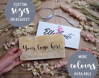 May include: Two wooden signs with custom text. The first sign is natural wood with the text "Your logo here". The second sign is white with the text "Elle  viste" and a pink dress illustration. The signs are on a wooden surface with dried flowers.