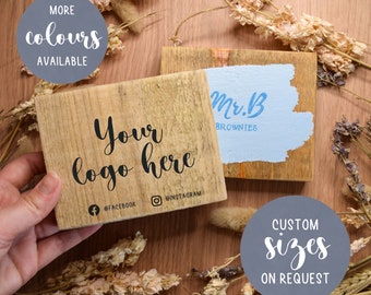 May include: Two wooden signs with custom text. The first sign says "Your logo here" with social media icons and the text "@facebook" and "@instagram". The second sign says "Mr. B Brownies" in blue paint on a light blue background. Both signs have a rustic, distressed look.