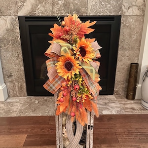 May include: A rectangular white wooden lantern decorated with a large arrangement of orange and yellow sunflowers, plaid ribbons in shades of green and brown, and artificial autumn leaves, berries, and foliage attached to the top section.