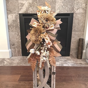 May include: A decorative lantern featuring a weathered gray wood frame with a central arched cutout. The top is adorned with a large arrangement of faux sunflowers, dried-look foliage, white blossoms, and layered ribbons in tan, olive, and cream.
