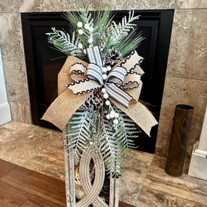 May include: A tall, rectangular wooden lantern with a distressed white finish, topped with a decorative arrangement of frosted pine branches, pinecones, white berries, and a multi-layered bow made of tan burlap and black and white striped fabric.