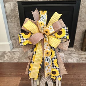 May include: A decorative bow crafted from multiple ribbons featuring sunflower patterns, yellow and white checkered prints, black and white buffalo plaid, and solid tan burlap. The bow has several loops and long trailing tails arranged in a cluster.