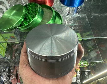 May include: A silver metal grinder is held in a hand, with red, blue, and green grinders nearby. The green grinder is open, showing its internal components. Glass smoking accessories are visible in the background, on a glass shelf.