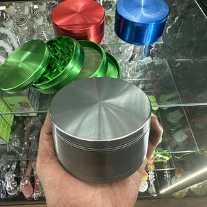 May include: A hand holds a silver metal grinder with a textured lid. Other grinders in red, blue, and green are displayed nearby. The green grinder is open, revealing its interior. Glass smoking accessories are visible in the background.