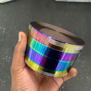 May include: A multi-coloured, rainbow-hued grinder. The cylindrical grinder is held in a hand and features a textured, knurled grip on the top and bottom. The colour transitions from yellow to blue to purple. The top is a dark, reflective colour.