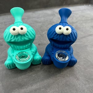 May include: Two novelty smoking pipes, one teal and one blue, shaped like a cartoon character with large white eyes. Each pipe has a cylindrical bowl and a curved mouthpiece. The pipes are made of a soft, molded material.