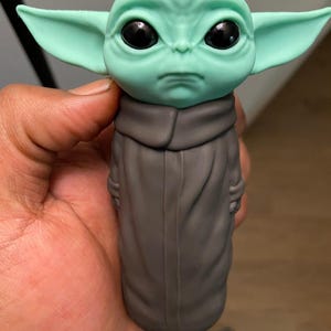 May include: A green and gray figurine of a character with large ears and black eyes. The figurine is wearing a gray robe. The character's head is green, and the ears are a lighter shade of green. The figurine is held in a hand.