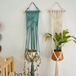 May include: Two macrame plant hangers, one teal and one white, hanging on a white wall. The teal hanger has a plant with green leaves in a white pot. The white hanger has a plant with green leaves in a brown pot.