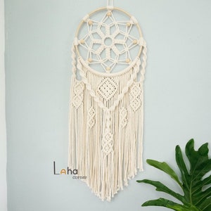 May include: A white macrame dream catcher with a wooden hoop and white beads. The dream catcher has a geometric pattern and is hanging on a light blue wall.