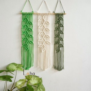 May include: Three macrame wall hangings with a leaf design. The hangings are made with natural cotton cord in green, white, and a darker green. Each hanging is attached to a wooden dowel.