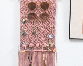 May include: A dusty rose macrame wall hanging organiser featuring a wooden dowel at the top with a hanging cord. The woven cotton cord design includes diamond patterns and fringe at the bottom, used to display sunglasses, earrings, and necklaces.
