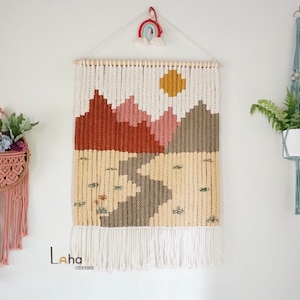 May include: A beige, brown, and pink macrame wall hanging with a white fringe. The hanging depicts a mountain range with a yellow sun in the sky. The hanging is labeled "Laha corner".