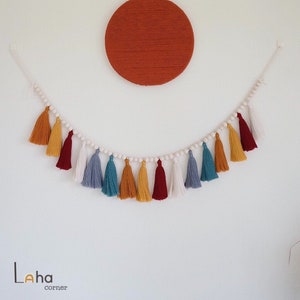 May include: A white string garland with wooden beads and colorful tassels in shades of red, yellow, blue, and white. The garland is hanging on a white wall with a large, round, orange wall hanging above it.