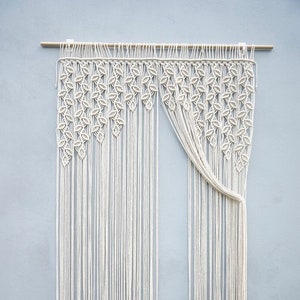 May include: A cream-colored macrame wall hanging suspended from a wooden rod. The upper section features a dense pattern of knotted leaf-like shapes, while the lower portion consists of long, vertical, unknotted fringe strands hanging straight down.