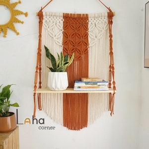 Könnte beinhalten: Ein Wandbehang aus Makramee mit einem zweistufigen Holzregal. Das Makramee ist aus weißem und orangefarbenem Garn gefertigt und hat ein geometrisches Muster. Das Regal hält eine Topfpflanze und einen Stapel Bücher. Der Text "Laha corner" ist in der linken unteren Ecke des Bildes sichtbar.