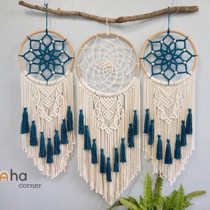 May include: A wall hanging featuring three circular wooden hoops suspended from a branch. The two outer hoops contain dark teal macrame patterns, while the center hoop features a white web design. All three include cream macrame fringe and teal tassels.