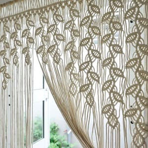 May include: An off-white macrame curtain hanging from a wooden rod, featuring a repeating leaf-like knot pattern across the top section that transitions into long, vertical, unknotted fringe strands that hang down to the bottom edge.