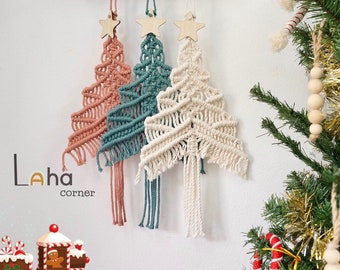 Könnte beinhalten: Drei Makramee-Weihnachtsbaum-Ornamente, die an einem Holzregal hängen. Die Bäume sind in den Farben Rosa, Türkis und Weiß gehalten. Jeder Baum hat einen Holzstern oben drauf.