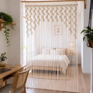 May include: A cream-colored macrame room divider featuring a series of knotted leaf-shaped patterns along the top edge, suspended from a horizontal wooden rod. The long, vertical cords hang down to create a textured, semi-transparent partition.