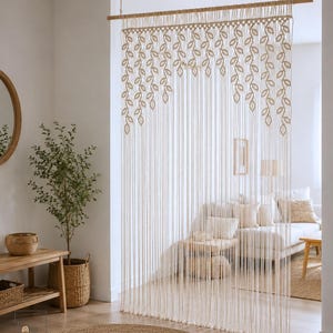 May include: A cream-colored macrame curtain hanging from a wooden rod, featuring a repeating leaf pattern at the top and long, vertical fringe strands that extend downward to create a decorative room divider with a knotted fiber design.