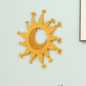 May include: A handmade, yellow macramé sunburst wall hanging with a crescent moon centre. The sun has radiating rays with tassel accents. A green cactus is in the foreground. The wall is painted a light blue.