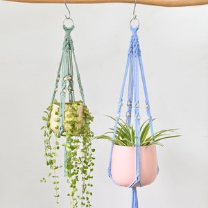 May include: Two macrame plant hangers with wooden beads, one is light blue and the other is light green. Both hangers are holding potted plants.