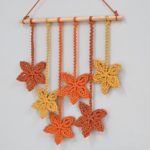 May include: A macramé wall hanging with seven flower-shaped ornaments in shades of orange and yellow. The flowers are suspended from a wooden dowel with a brown cord. A houseplant in a brown pot sits below.