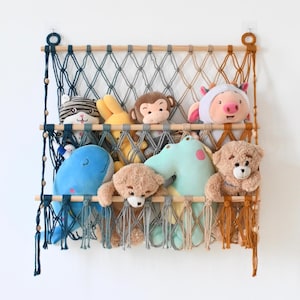 May include: A wall-mounted macrame storage hammock featuring three horizontal wooden dowels and a knotted cord net in an ombre color gradient from dark blue to orange. The net holds various plush toys including a blue whale and teddy bears.