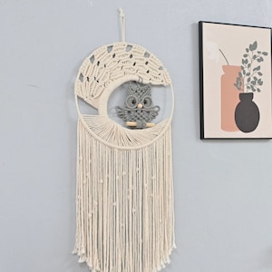 Könnte beinhalten: Cremefarbener Makramee-Wandbehang mit einem Baumdesign und einer grauen Eule, die auf einem Holzast sitzt. Die Eule hat große Augen und eine detaillierte Textur. Der Wandbehang hat lange, herabhängende Fransen.