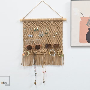 Peut inclure: Une suspension murale en macramé beige avec une tige en bois et une corde pour la fixation. Le tissage en grille supporte diverses boucles d'oreilles, deux paires de lunettes, des élastiques à cheveux et deux colliers perlés, avec des franges.