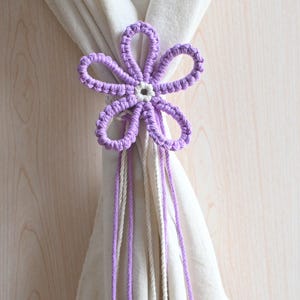 May include: A handcrafted, flower-shaped curtain tieback in shades of lavender and white. The macrame design features a central flower with six petals and dangling cords. The tieback is shown holding a white curtain against a light wood background.