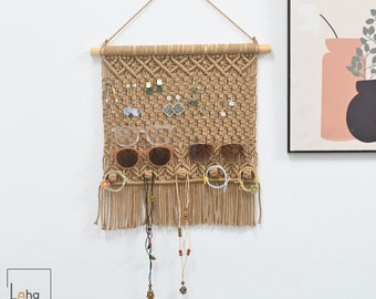 May include: A tan macrame wall hanging organiser featuring a wooden dowel and cord for suspension. The woven grid holds various earrings, two pairs of glasses, hair ties, and two beaded necklaces, with a fringe finish at the bottom edge.
