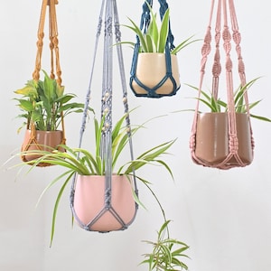 Puede incluir: Cuatro maceteros colgantes de macramé con cordones de diferentes colores y cuentas de madera. Los maceteros sostienen plantas en macetas. Los maceteros están colgados de una barra de madera.