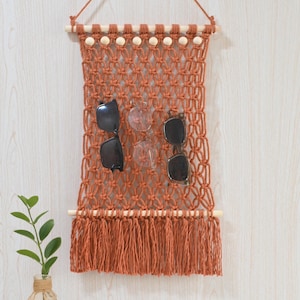 May include: A terracotta-colored macrame wall hanging featuring a knotted net pattern, wooden dowels, and light-colored wooden beads. The piece includes a fringe at the bottom and holds two pairs of sunglasses. The text Laha corner is visible.
