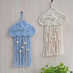Puede incluir: Dos colgantes de pared de macramé con forma de nube. Uno es azul claro, el otro blanco roto. Ambos tienen una forma de nube anudada en la parte superior con hebras colgantes y cuentas de madera. Se muestran sobre un fondo de madera clara.
