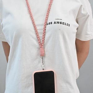 May include: A pink braided lanyard with a matching phone case. The phone case is attached to the lanyard with a silver clasp. The white t-shirt has the words "California LOS ANGELES" printed on the front.
