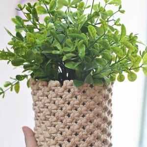 May include: A small, woven macrame plant pot in a natural beige color, filled with vibrant green artificial foliage. The pot has a textured, open weave design. The plant is held in a hand, with a blurred background.