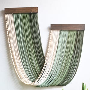 May include: A wall hanging featuring two dark brown wooden bars from which long, draped strands of cream and sage green cord are suspended. A single string of small, round, light-coloured wooden beads follows the curve of the cord arrangement.
