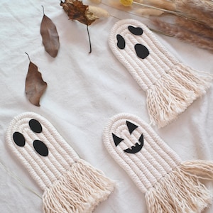 May include: Four handmade macrame ghost decorations with black felt eyes and mouths. The ghosts are white with a fringed bottom. The ghosts are arranged on a white surface with dried leaves and grasses.