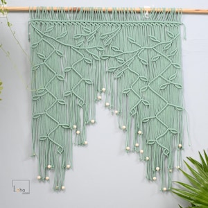 May include: A sage green macrame wall hanging featuring an intricate vine and leaf pattern knotted from cotton cord. The design includes long, flowing fringe strands accented with small, round, light-colored wooden beads at the bottom ends.