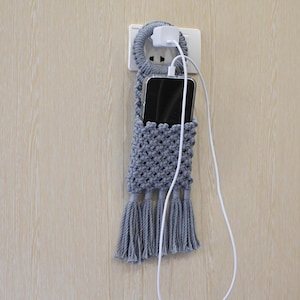 May include: A gray macrame phone holder with a tassel fringe, hanging from a wall outlet. A white charging cable is plugged into the outlet and connected to a black smartphone inside the holder. The holder is made of woven cotton.