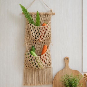 Puede incluir: Un tapiz de macramé beige con dos bolsillos abiertos suspendidos de un listón de madera. Los bolsillos contienen hojas de helecho verdes, un pepino verde y zanahorias naranjas. La parte inferior presenta flecos largos y rectos.