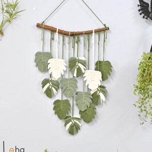 May include: A macrame wall hanging with green and white leaves. The leaves are made of yarn and are hanging from a wooden branch. The wall hanging is on a light gray wall.