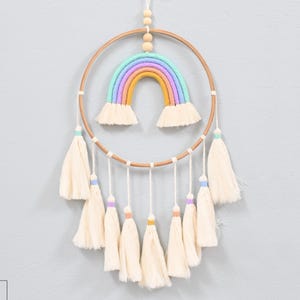 May include: A handmade rainbow dreamcatcher with a wooden hoop and cream-colored tassels. The rainbow features pastel colors and is suspended from a white cord with wooden beads. The dreamcatcher is designed for wall decor.