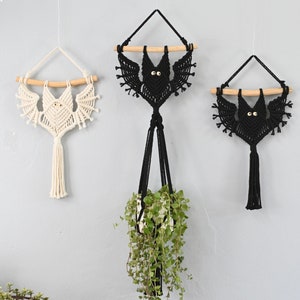May include: Three macrame bat wall hangings, one white, one black, and one black with a green plant hanging from it.