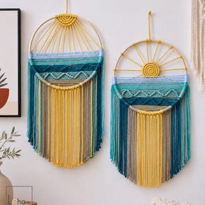 May include: Two circular macrame wall hangings featuring wooden hoops, yellow sun-like patterns, and layered horizontal bands in shades of teal, light blue, and beige. The designs include intricate knotted textures and long, flowing fringe.