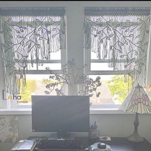 May include: Two dark gray macrame window valances featuring an intricate leaf and vine pattern with long, dangling fringe. The curtains are hung over two windows, displaying a knotted cord design that creates a decorative, textured appearance.