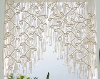 May include: An off-white macrame wall hanging featuring a leaf and vine pattern suspended from a wooden dowel. The intricate knotted cord design creates a cascading effect over a window. The text Laha corner is visible in the bottom left corner.