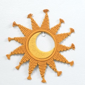 Könnte beinhalten: Ein kreisförmiger Makramee-Wandbehang mit einem senfgelben Sonnenstrahlen-Design. Die strahlenförmigen Spitzen sind jeweils mit einer kleinen Quaste versehen. In der Mitte befindet sich eine hellgelbe Mondsichel aus texturierter Kordel.