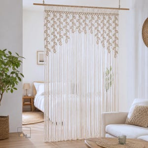 May include: A cream-colored macrame room divider hanging from a wooden rod. The top section features a repeating pattern of knotted leaf shapes, while the lower portion consists of long, vertical, unknotted fringe strands that hang straight down.